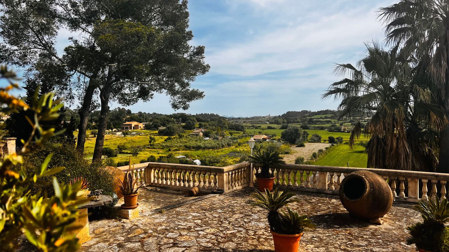 Scenic Mediterranean villa terrace with stone balustrade, lush gardens, palm trees, and panoramic countryside views under blue sky.