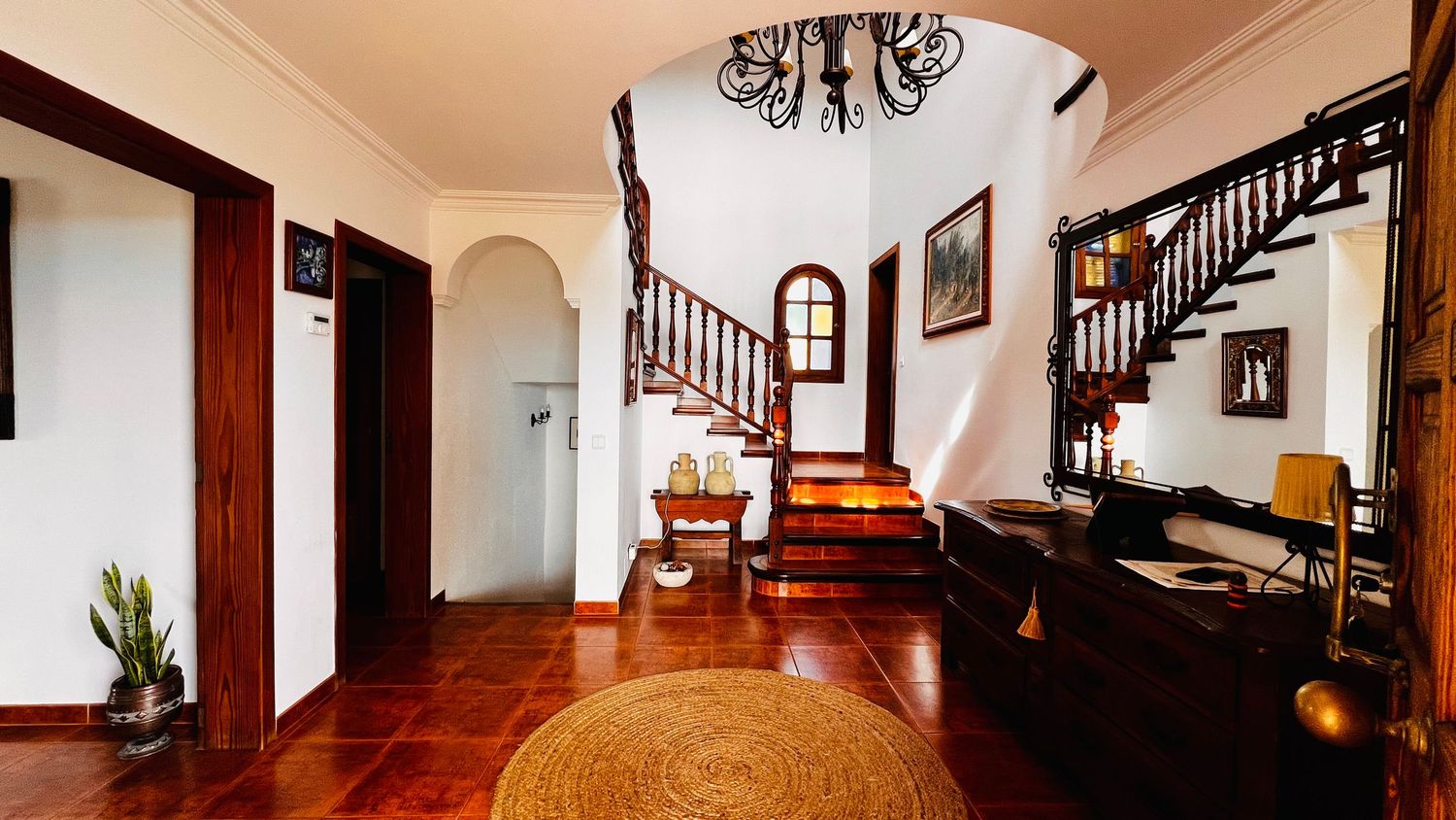 Elegant home entryway with terracotta tile floors, ornate wooden staircase, arched doorways, and a decorative chandelier.