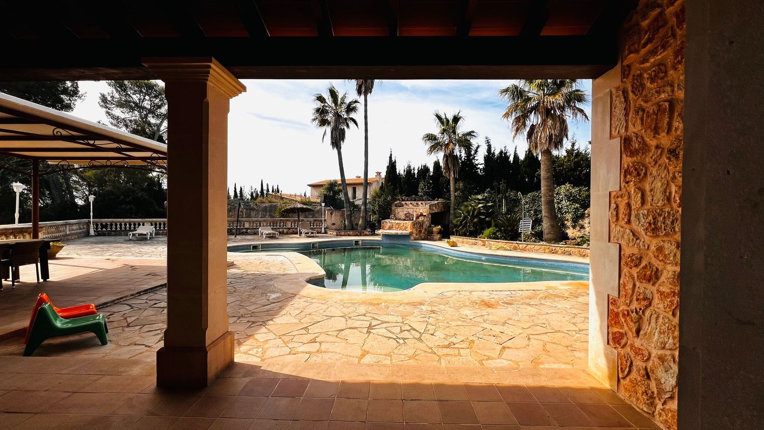 Stone-pillared covered terrace overlooking a sparkling outdoor pool surrounded by palm trees in a Mediterranean villa.