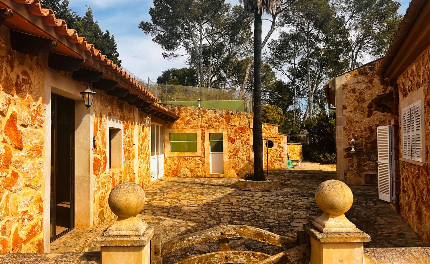Rustic stone courtyard with terracotta roof buildings, cobblestone path, and lush trees in warm golden sunlight.