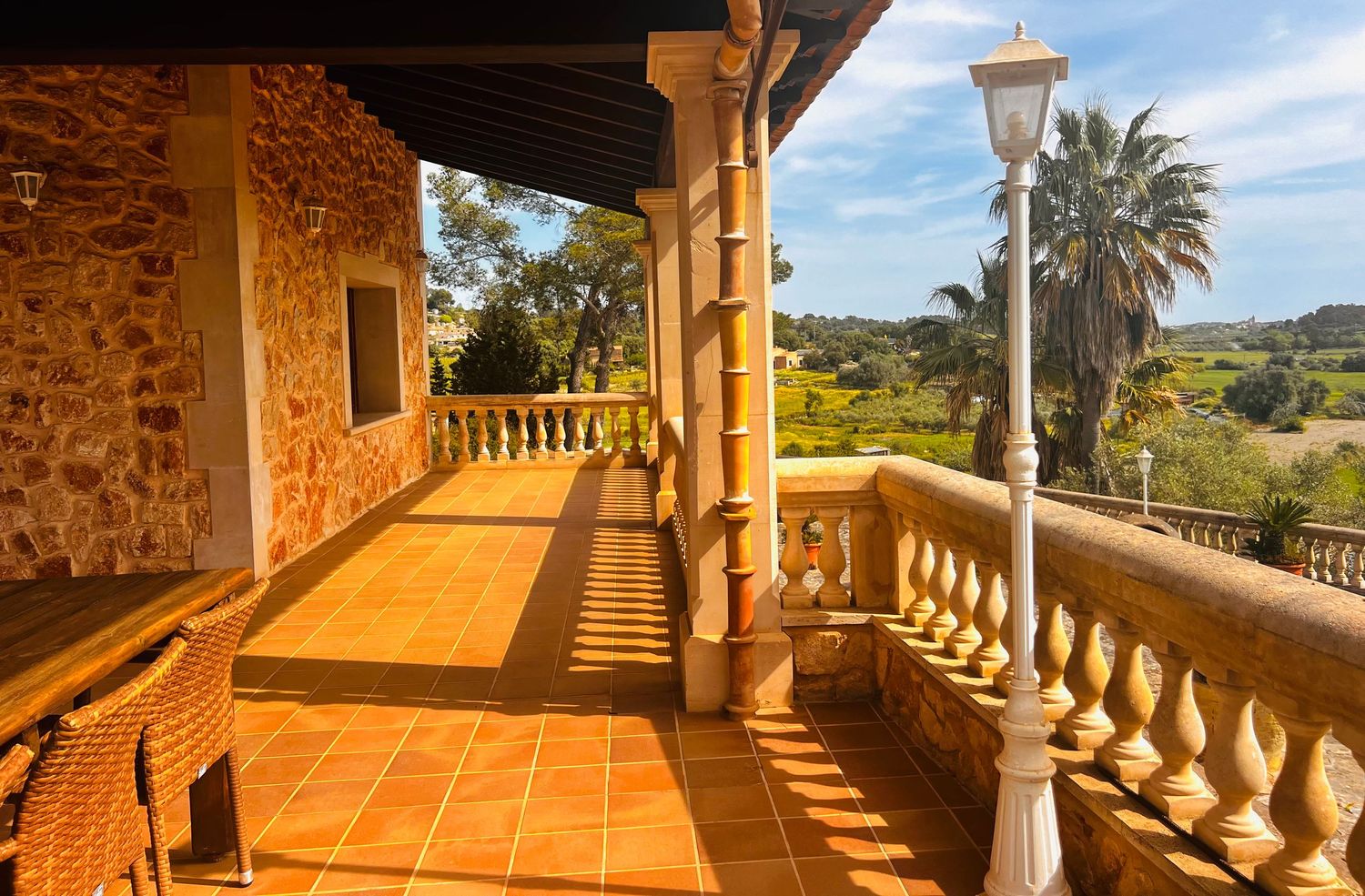 Sunlit terracotta-tiled balcony of a rustic stone villa with white balustrades, a lamp post, and lush green landscape views.