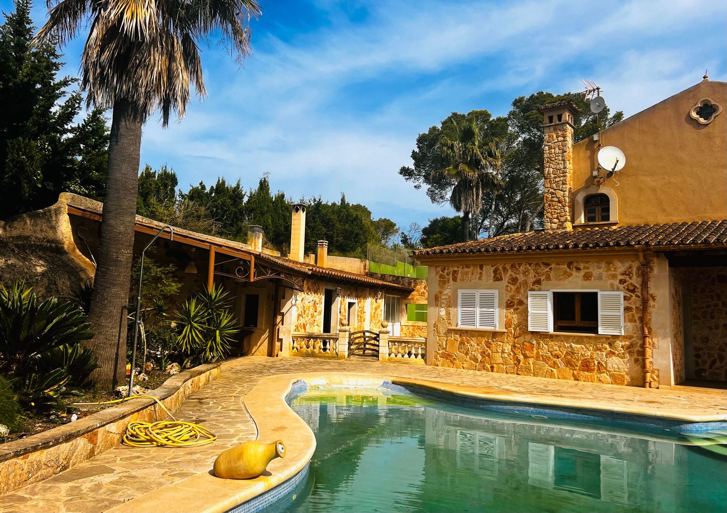 Traditional stone finca with terracotta roof beside a serene outdoor pool surrounded by lush palm trees in Mallorca.