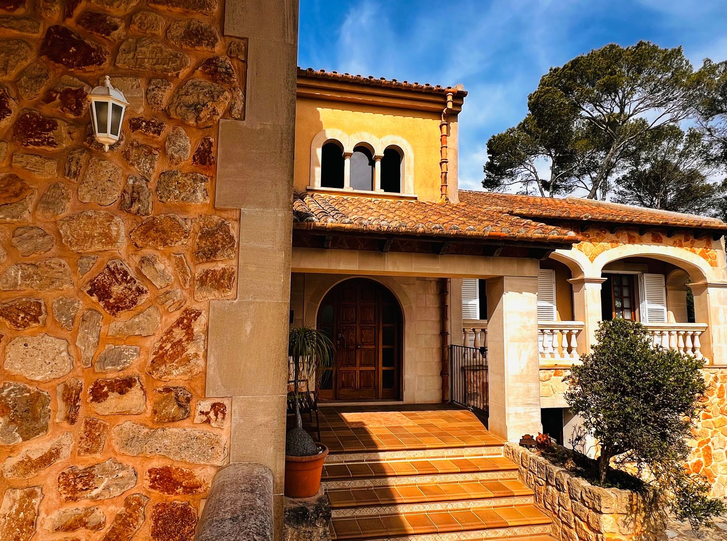 Elegant Mediterranean villa with stone facade, arched windows, terracotta roof, and wooden entrance door under a sunny blue sky.