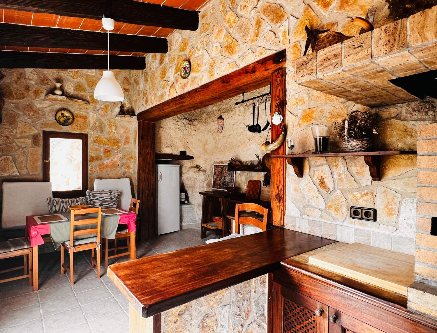 Rustic stone-walled kitchen and dining area with wooden furniture, hanging pots, and warm Mediterranean interior design.