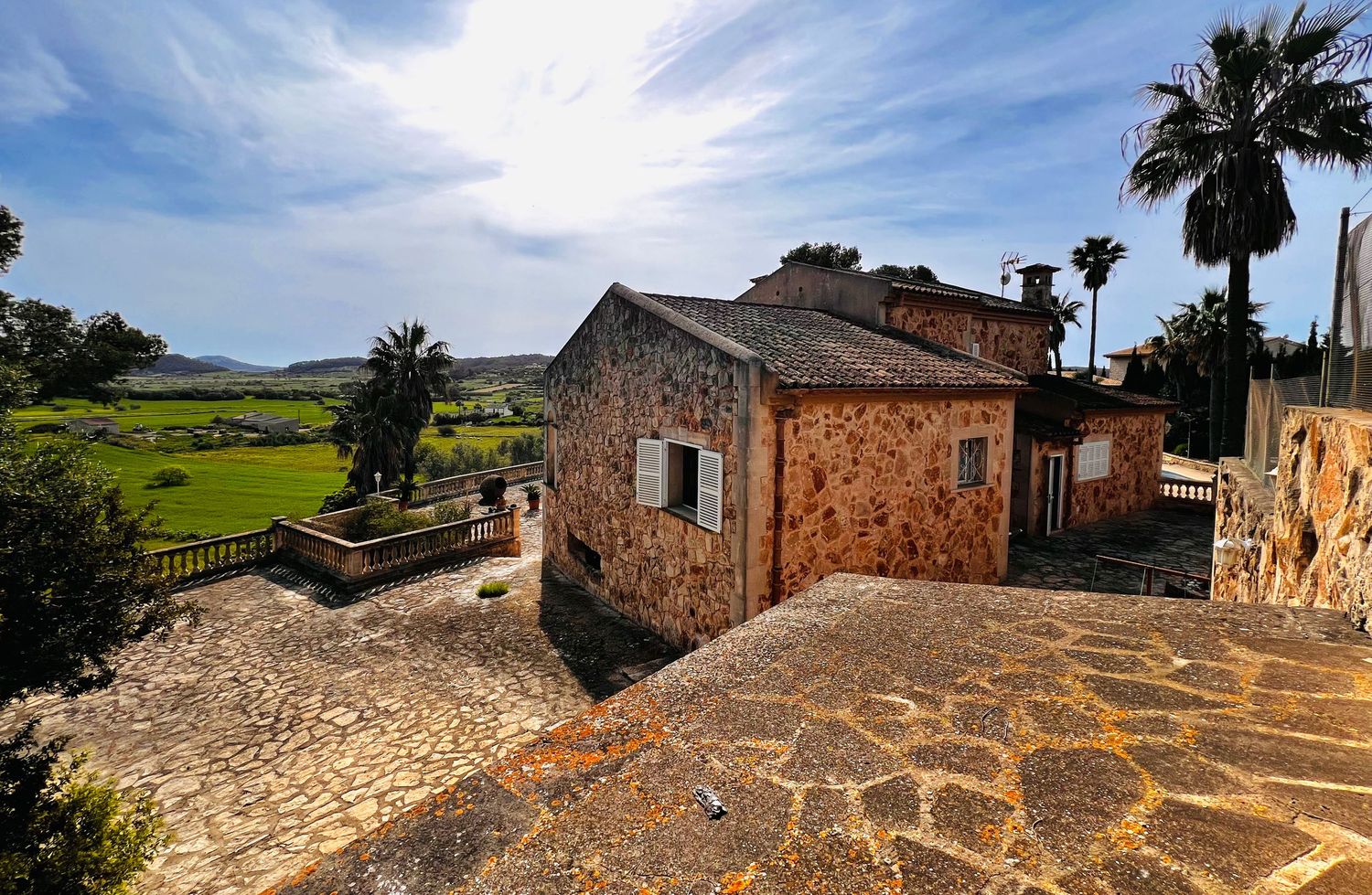 Stone villa with terracotta roof overlooking lush green fields, palm trees, and mountains under a blue sky in Mallorca.