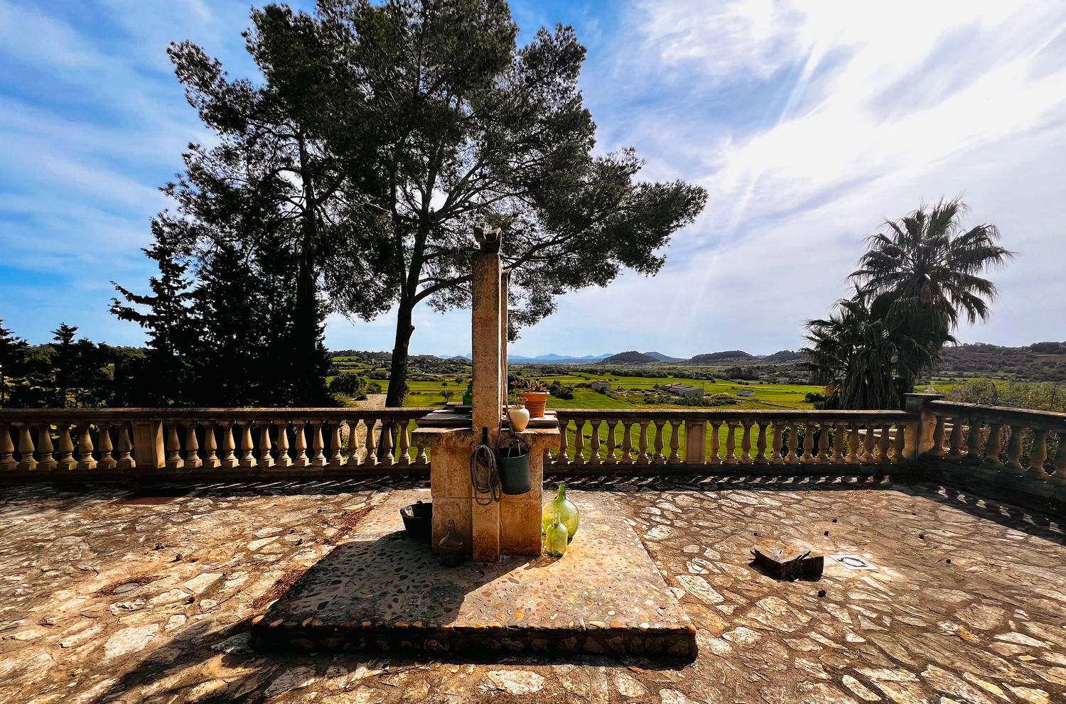 Stone monument on a terrace with balustrade, surrounded by trees and scenic countryside views under a partly cloudy sky.