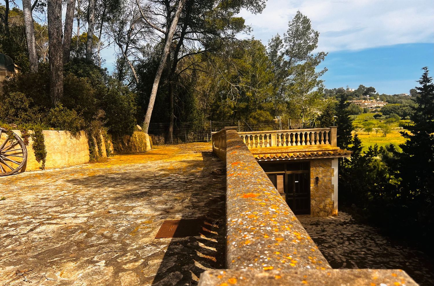 Stone terrace with balustrade overlooking lush gardens and countryside, surrounded by tall trees under a bright blue sky.