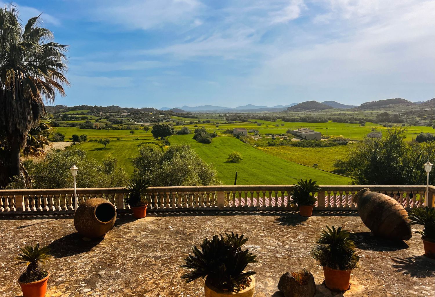 Panoramic view from a stone terrace with potted plants overlooking lush green valley and rolling hills under blue sky.