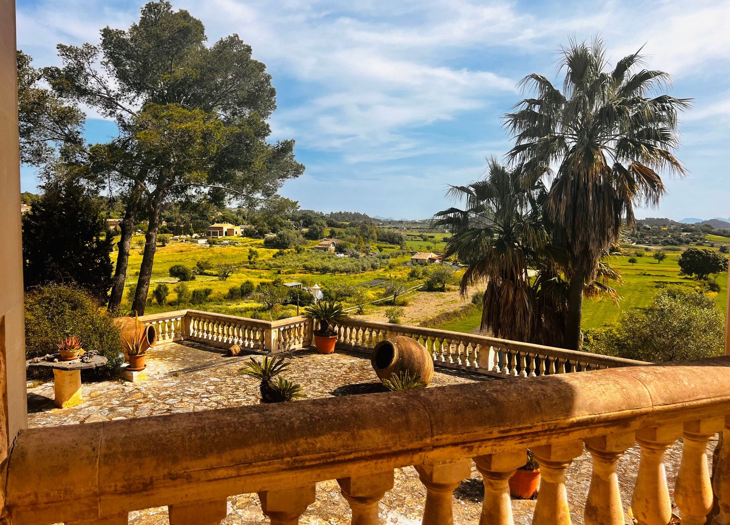 Stone balcony with ornate balusters overlooking lush green countryside with palm trees under a clear blue sky.