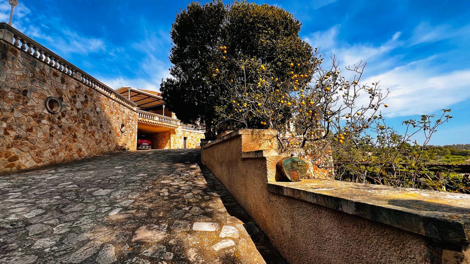 Stone villa with scenic driveway, terrace wall, and large tree under a vivid blue sky.