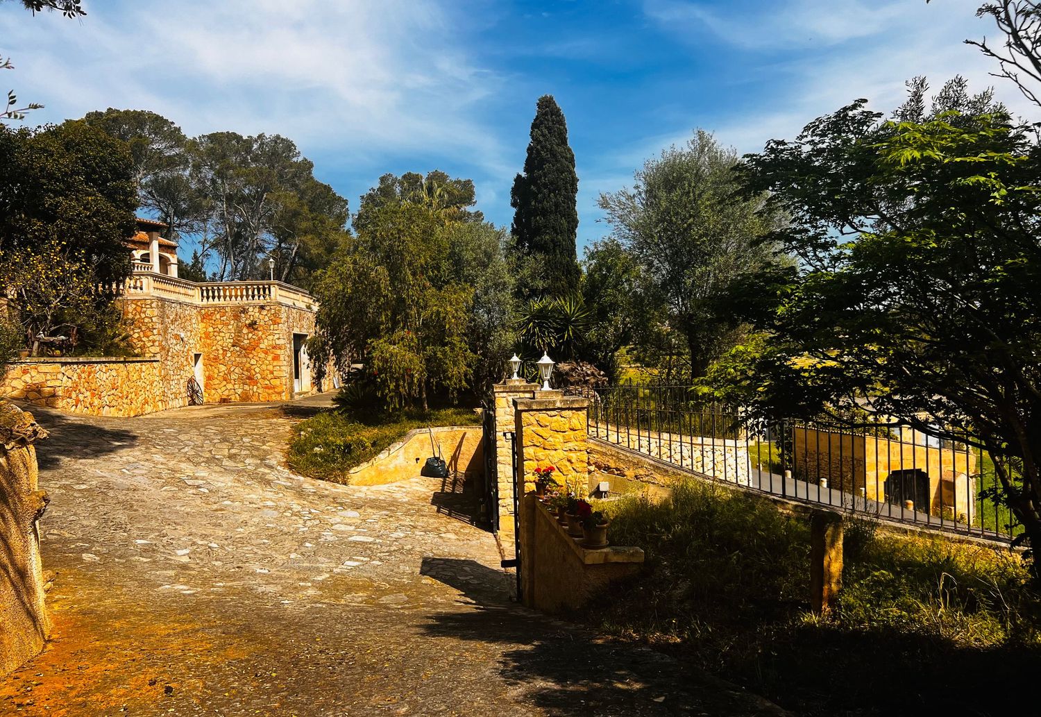 Historic stone garden path with ornate white fence, cypress trees, and rustic architecture under a blue sky.