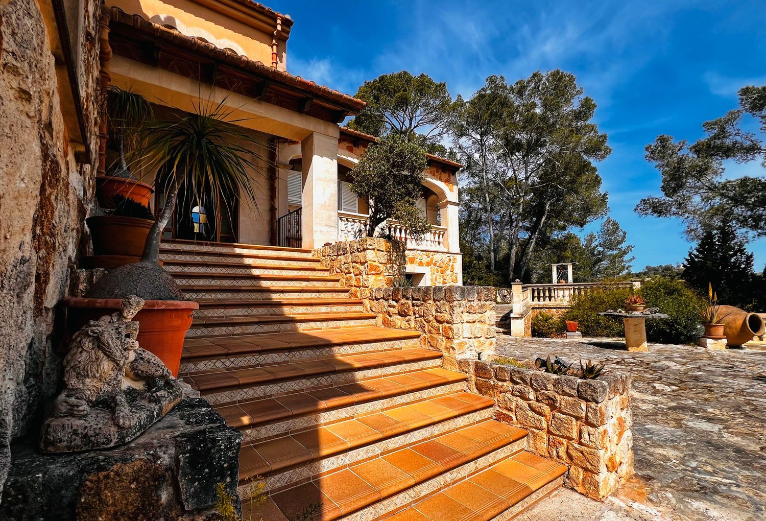 Elegant stone villa with wooden staircase, terracotta pots, and lush trees under a bright blue sky.