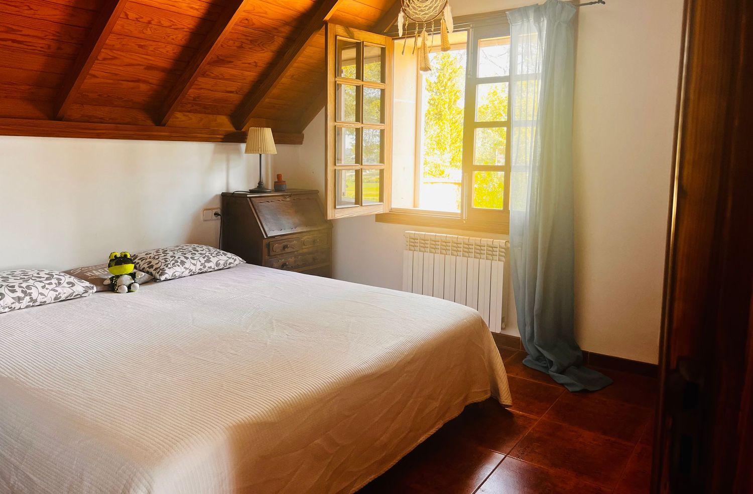 Cozy attic bedroom with wooden ceiling, white bedding, hardwood floors, and a window with sheer curtains letting in natural light.