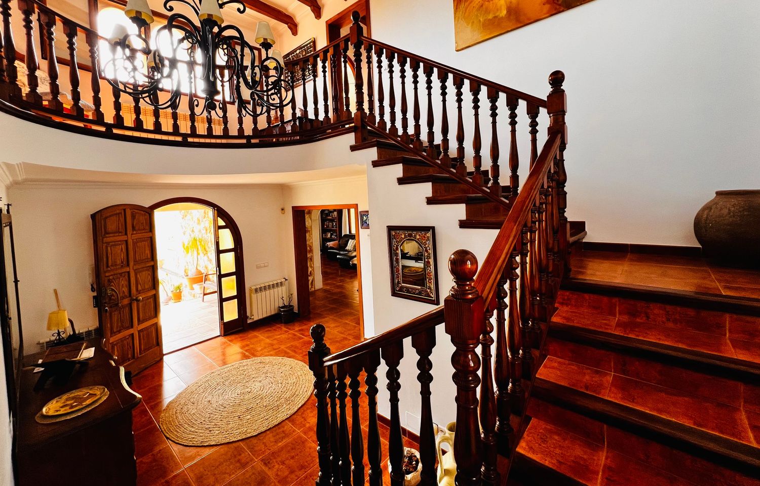 Elegant two-story foyer with dark wood staircase, wrought iron chandelier, arched doorway, and terracotta tile floors.