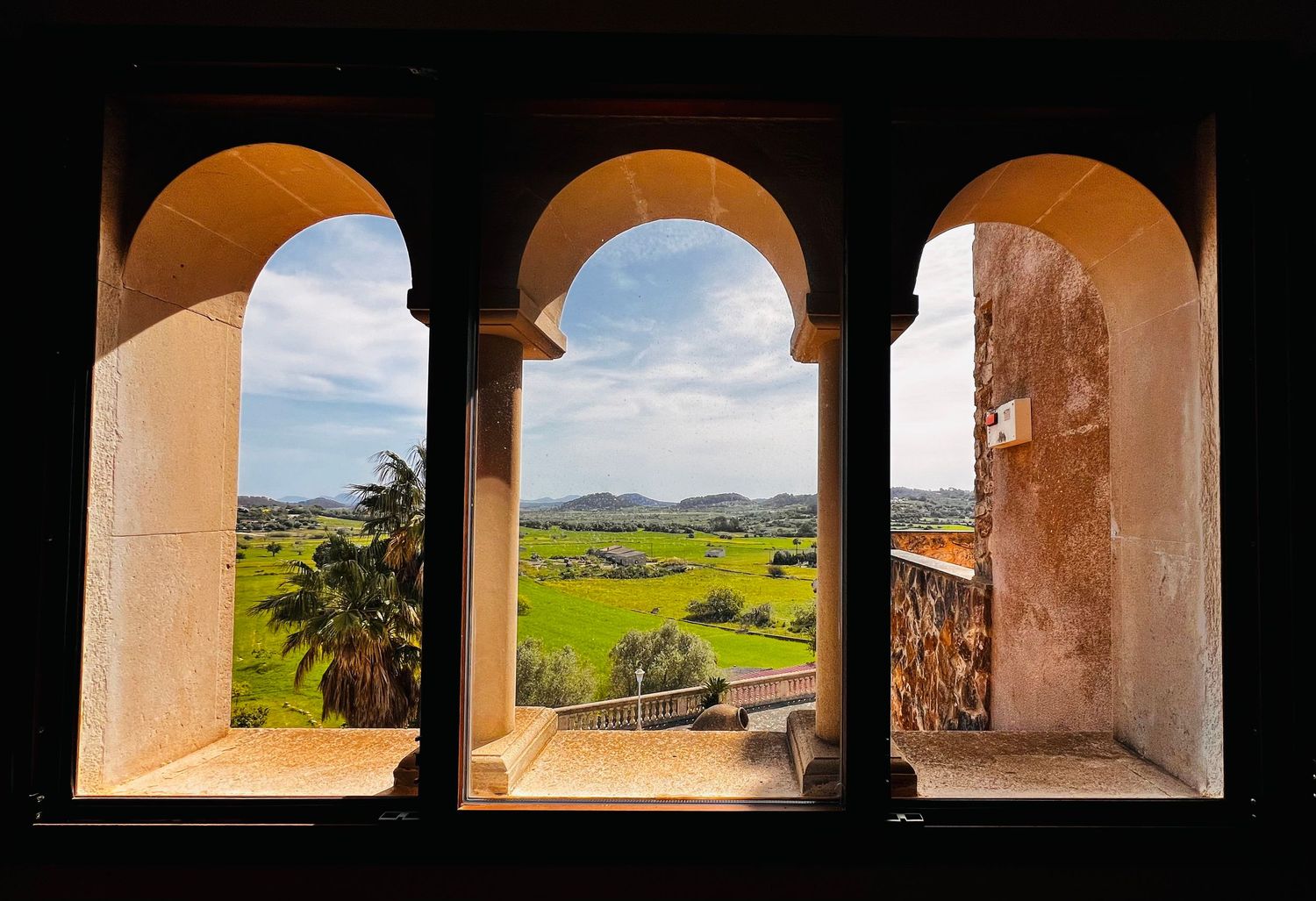 Three arched stone windows frame a scenic view of lush green fields, palm trees, and rolling hills under a blue sky.