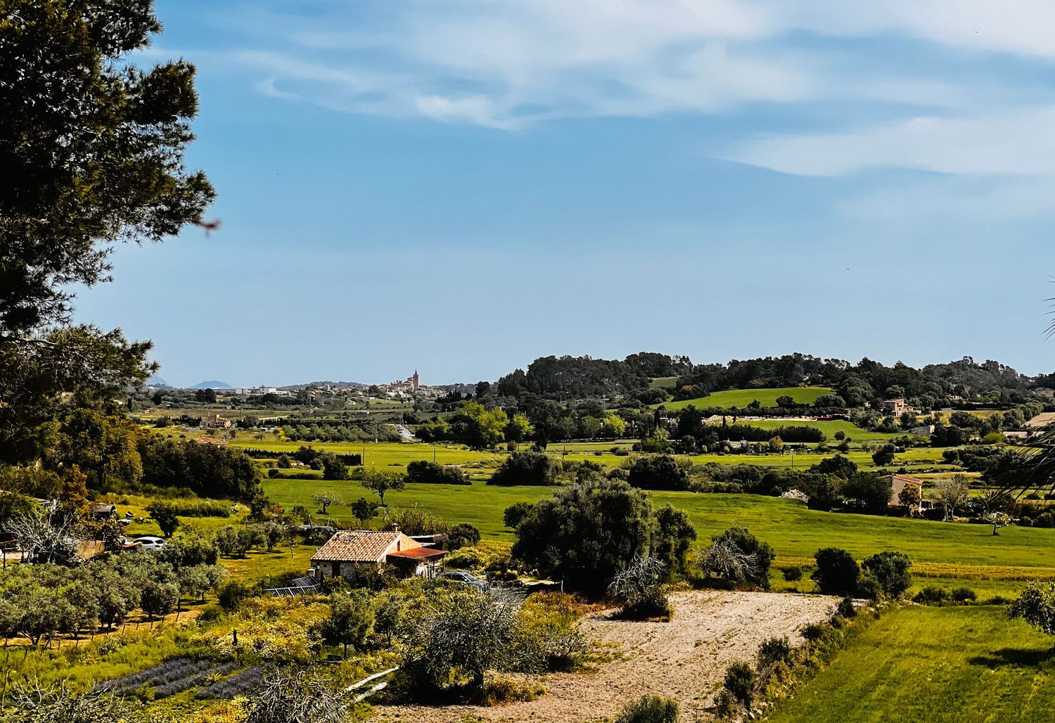 Panoramic view of lush green countryside with farmhouses, rolling hills, and blue sky in a Mediterranean rural landscape.