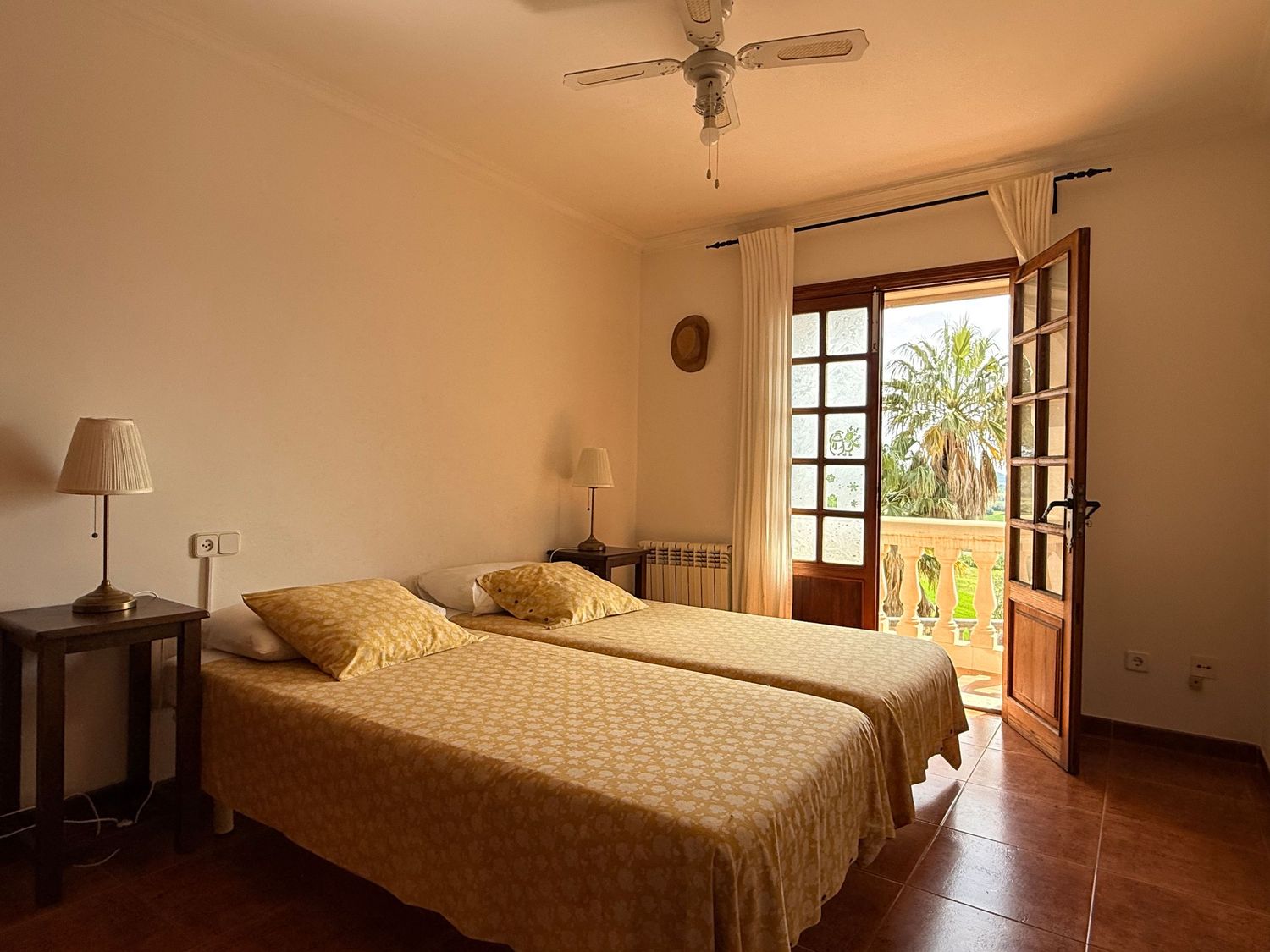Bright twin bedroom with yellow bedding, terracotta floors, ceiling fan, and wooden balcony doors overlooking a palm tree.