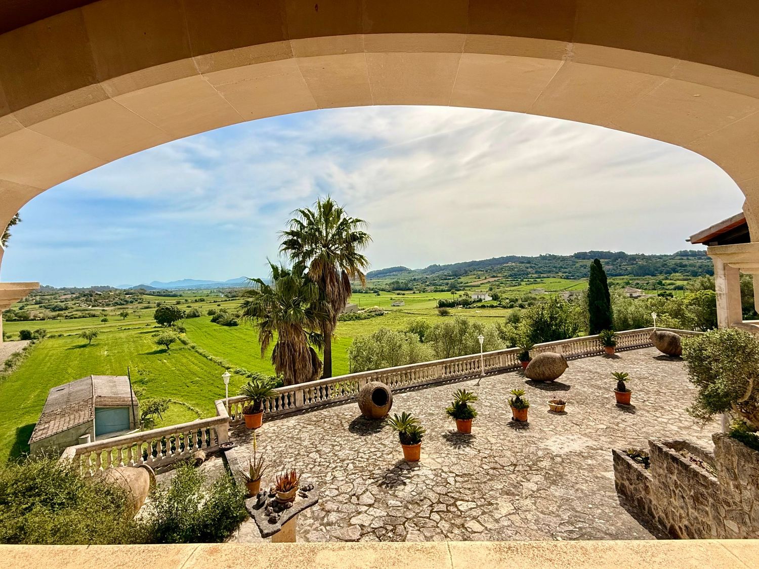 Stone archway terrace overlooking lush green valley with palm tree, potted plants, and mountain views under blue sky.