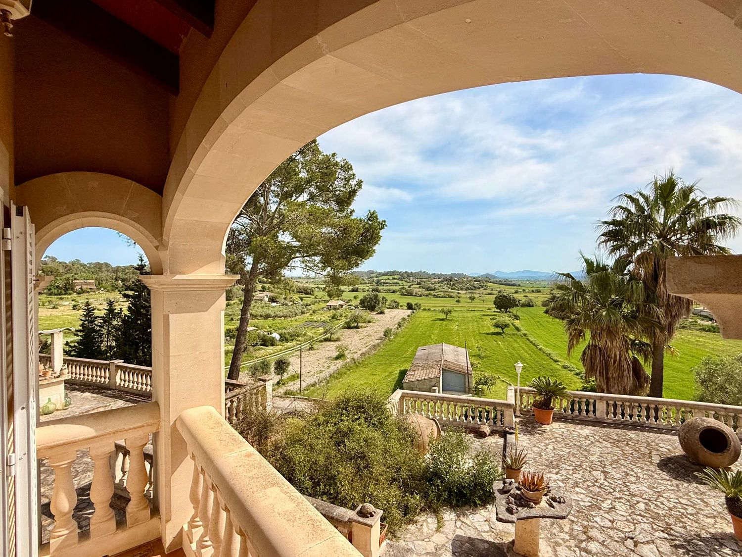 Elegant arched balcony overlooking lush green gardens and rolling countryside with palm trees and blue sky in Mallorca.