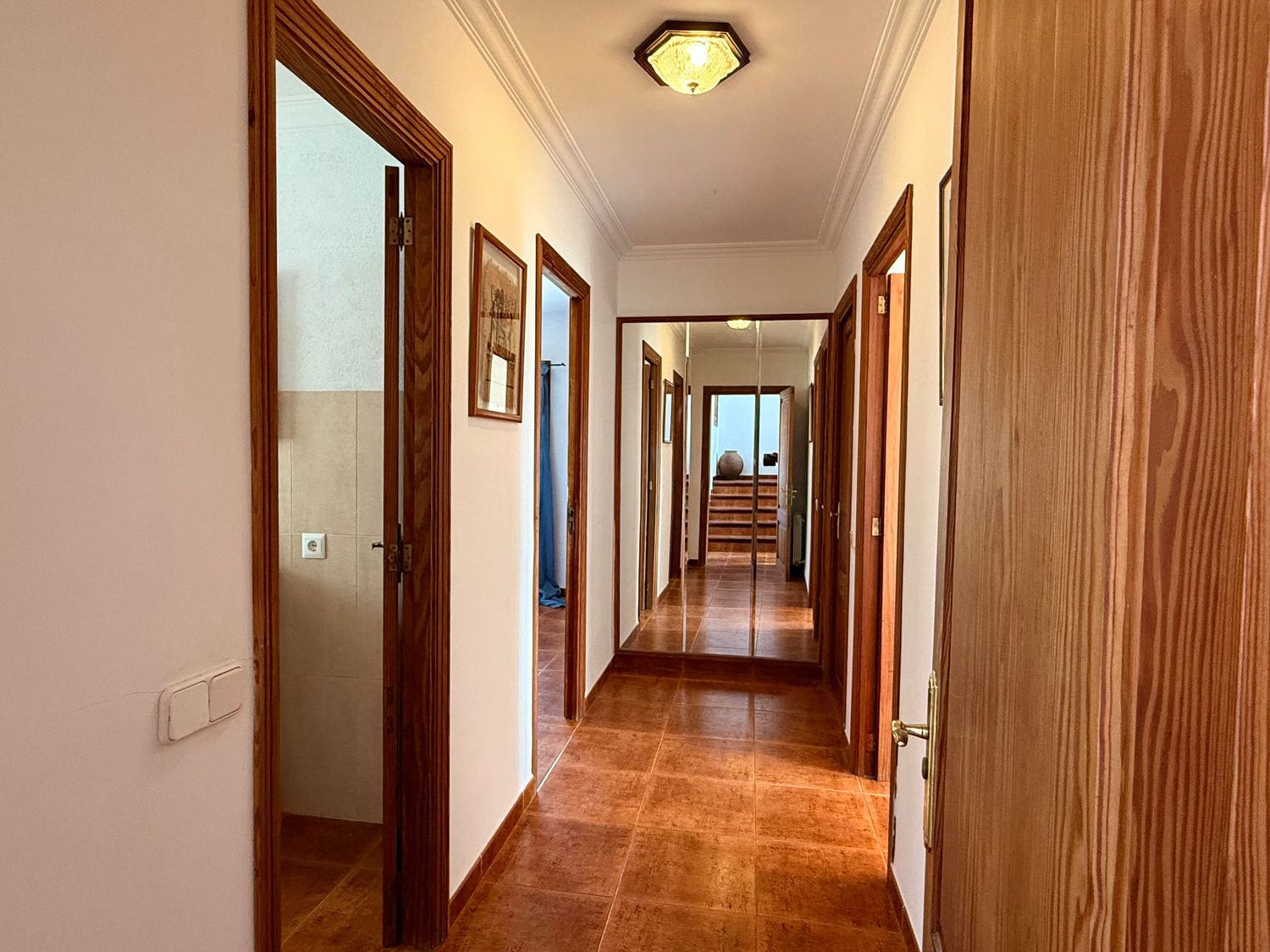 Long hallway with terracotta tile floors, wooden doors, and white walls leading to a bright window at the end.