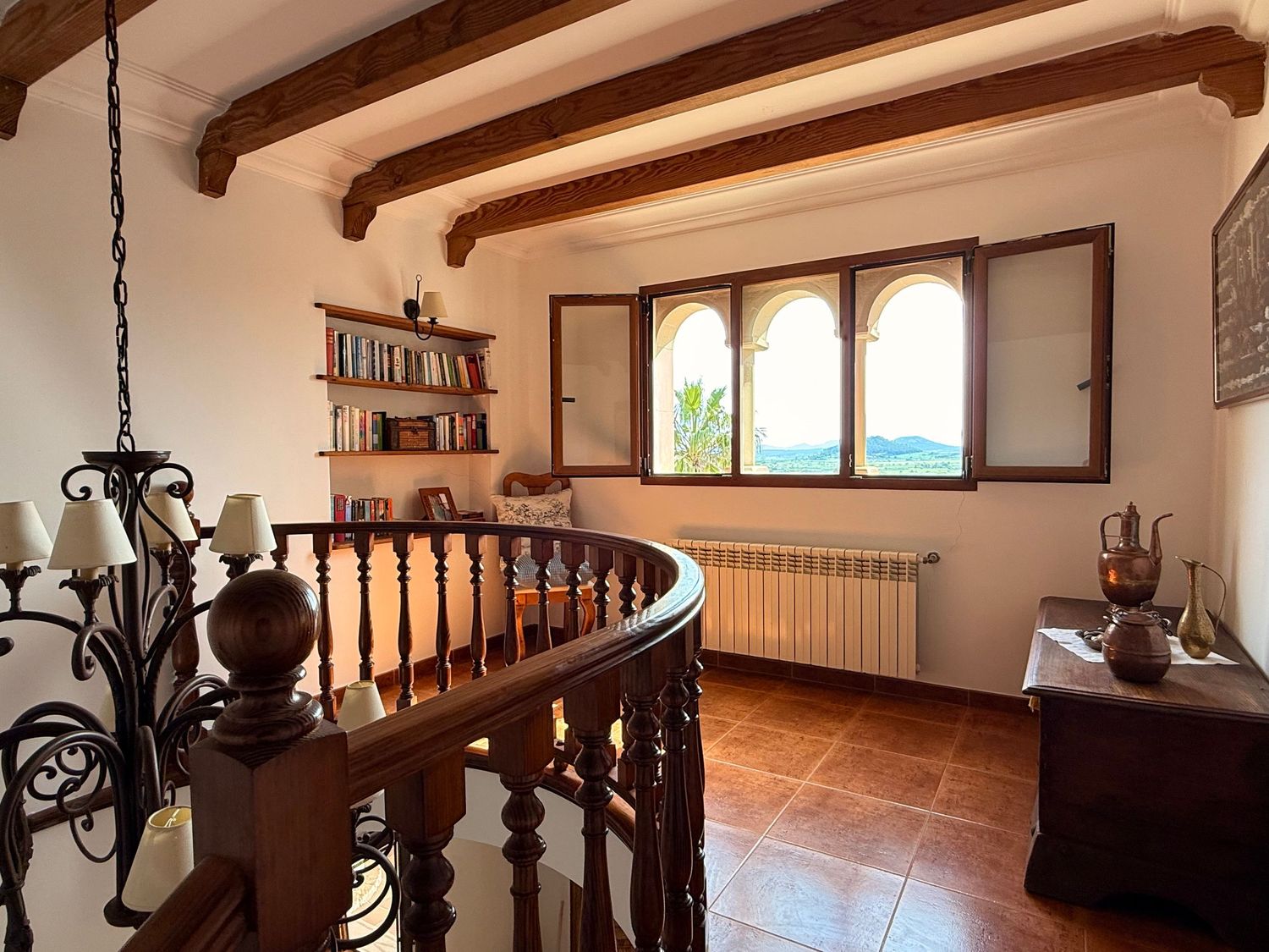 Rustic interior landing with wooden beamed ceiling, curved banister, arched windows, terracotta tiles, and a decorative chandelier.