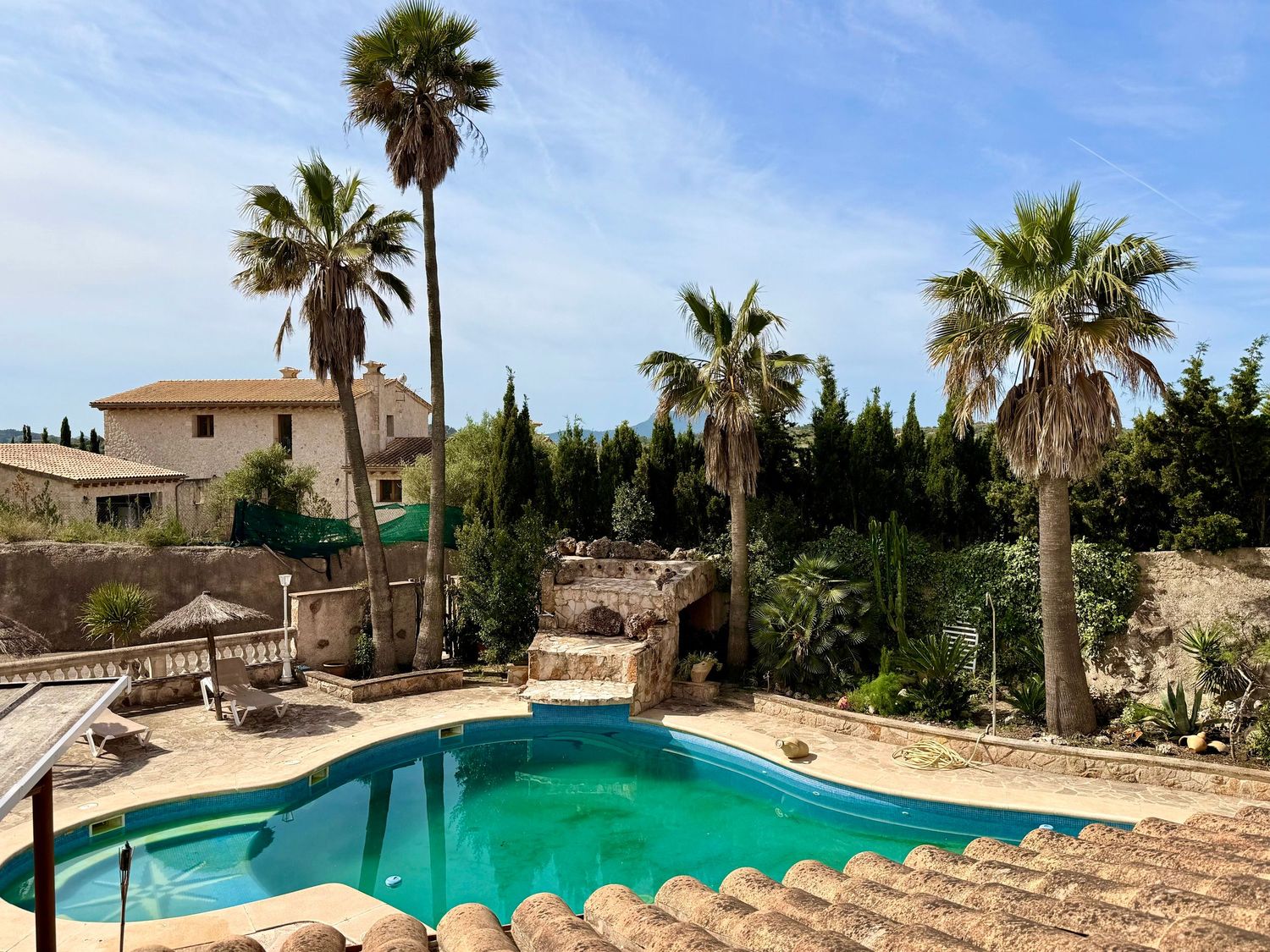 Outdoor pool surrounded by palm trees and stone landscaping under a clear blue sky at a Mediterranean villa.