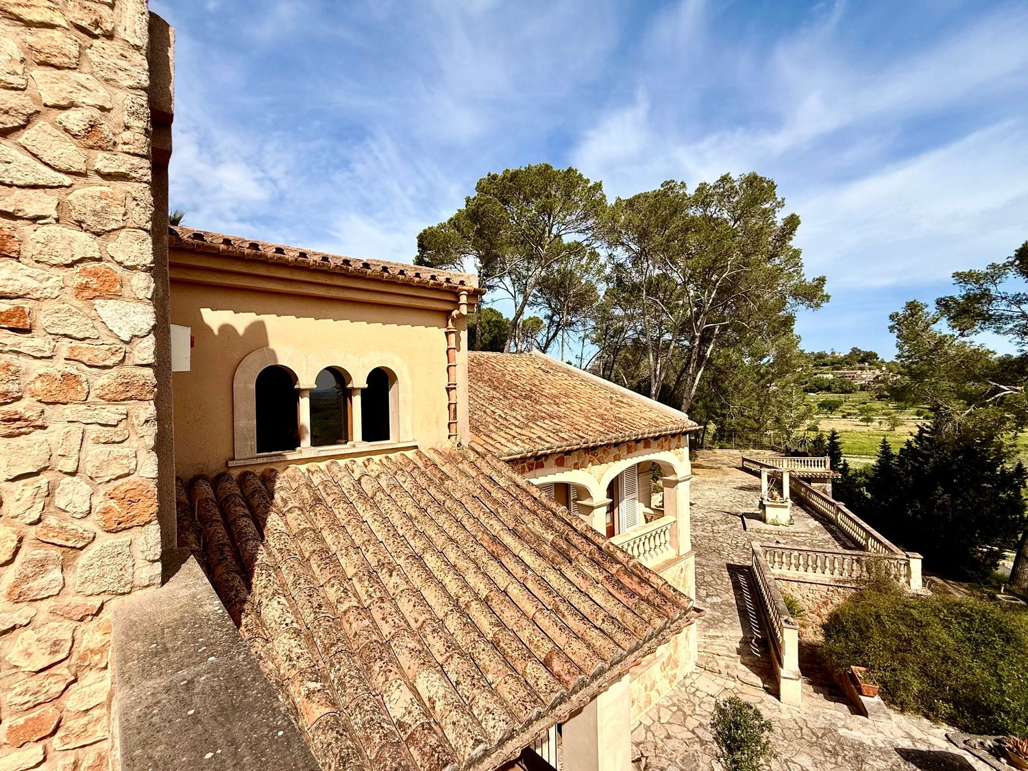 Rustic Mediterranean villa exterior with terracotta roof tiles, stone walls, arched windows, and lush green trees under a blue sky.