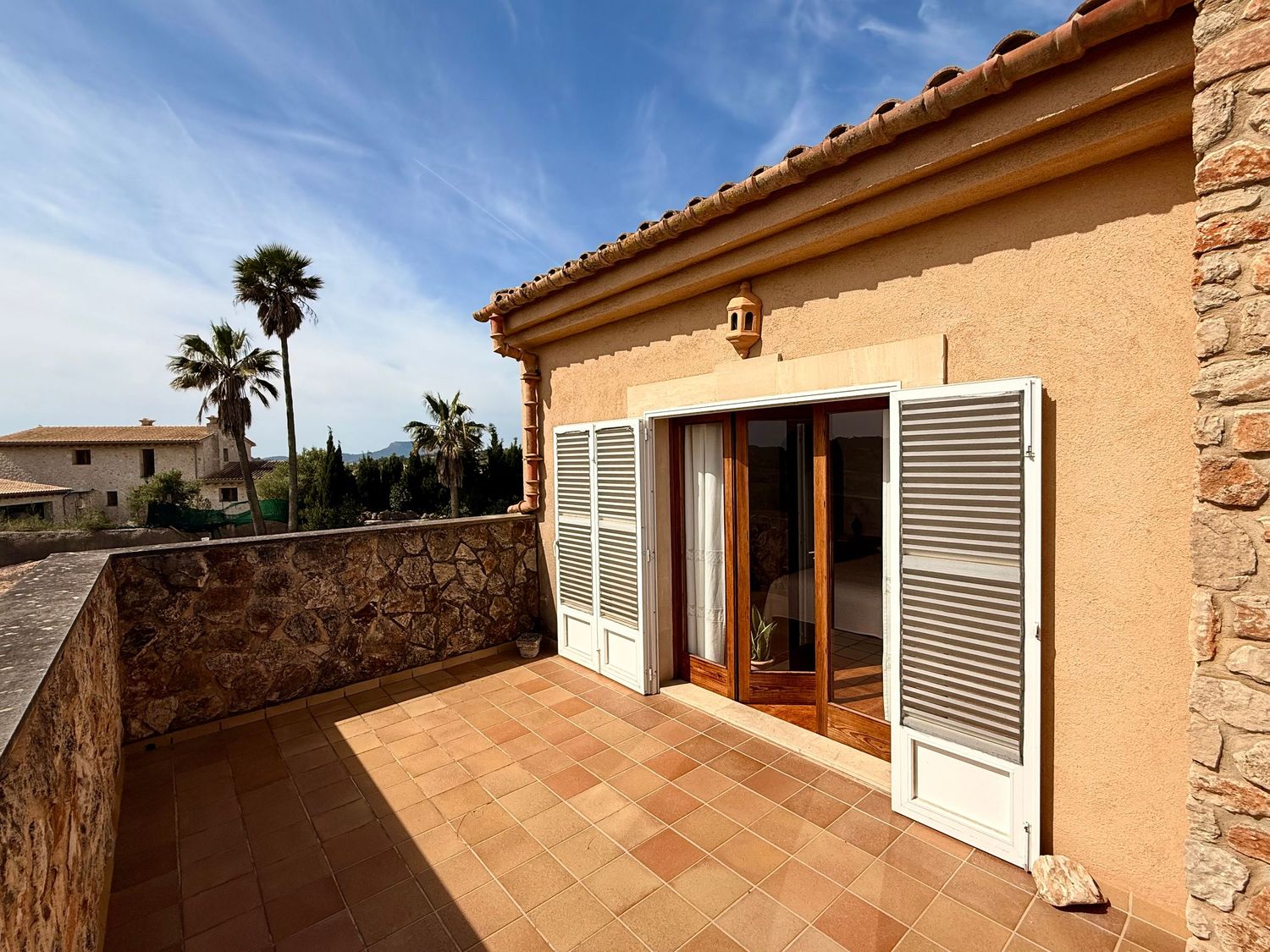 Sunny terrace of a Mediterranean villa with terracotta tiles, white shuttered doors, and stone wall views.