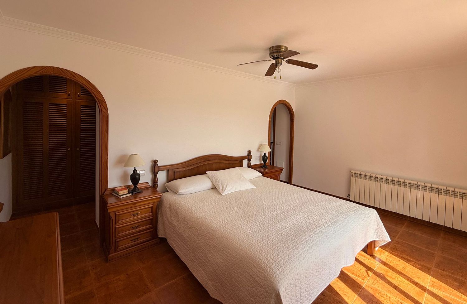 Spacious bedroom with white bedding, wooden furniture, arched doorways, ceiling fan, and parquet flooring in a Mediterranean villa.
