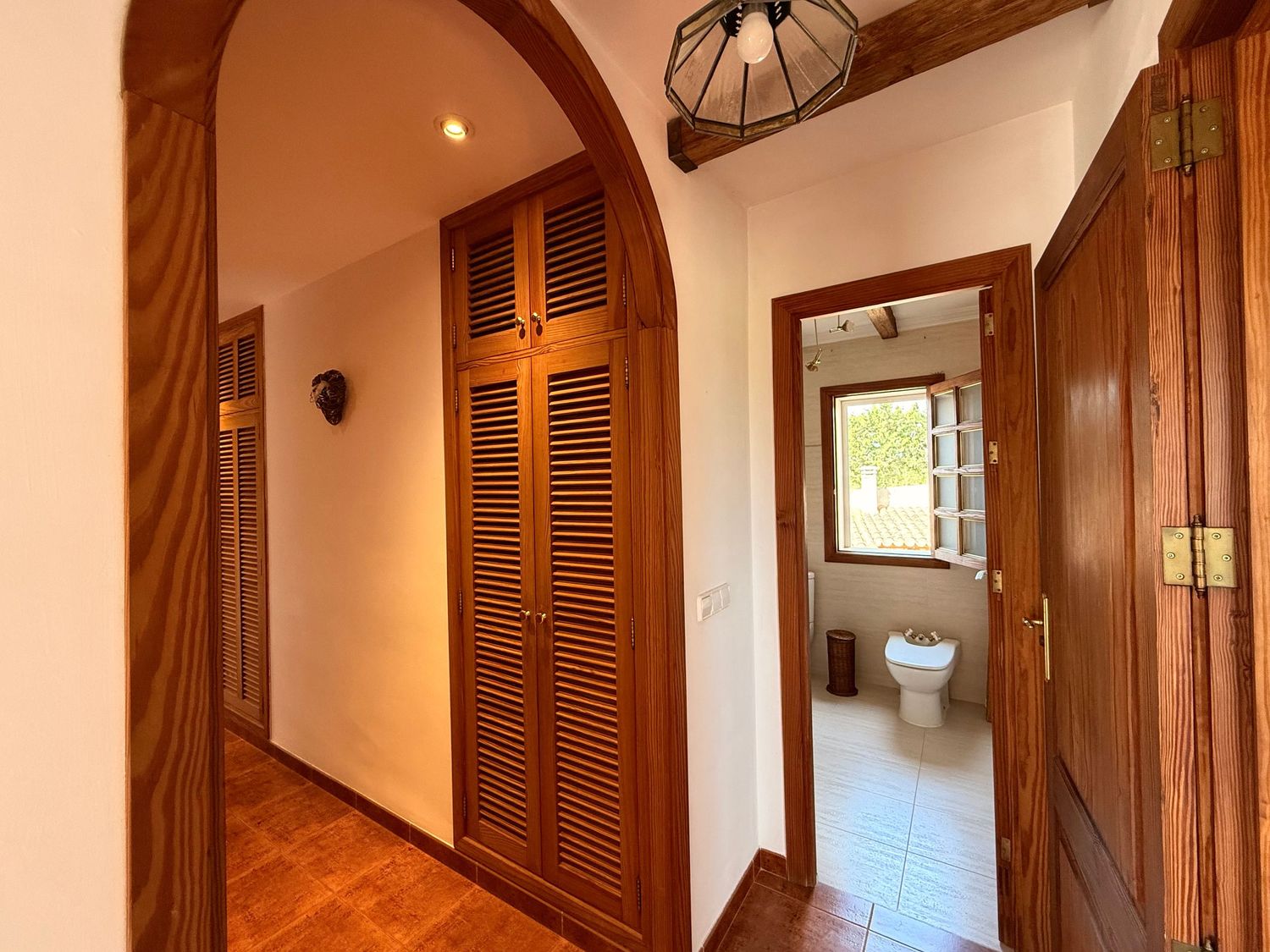 Elegant interior hallway with wooden louvered doors, arched doorway, terracotta tile floors, and a view into a bright bathroom.