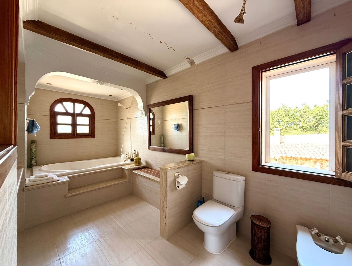 Elegant bathroom with beige tiles, arched window over bathtub, wooden ceiling beams, toilet, and bidet in natural light.