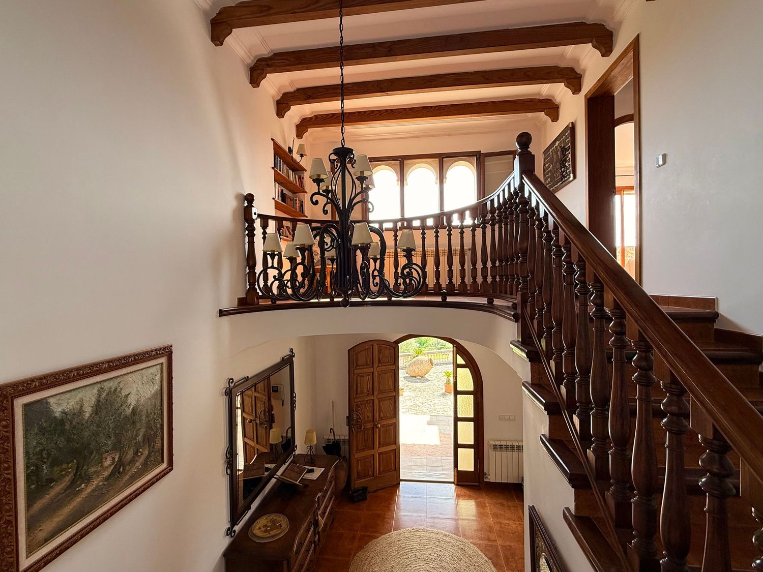 Elegant foyer with dark wood staircase, wrought iron railing, terracotta tile floors, and arched doorway leading to a sunlit garden.