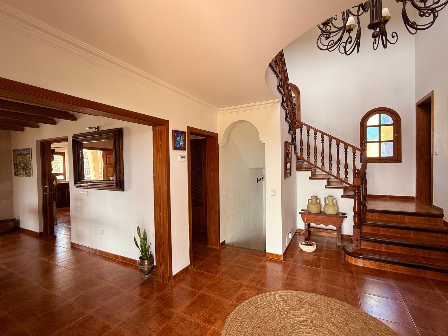 Spacious villa hallway with terracotta tile floors, elegant wooden staircase, arched doorways, and warm neutral walls.