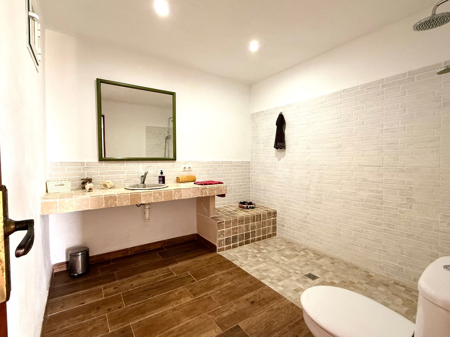 Modern bathroom with wood-look tile floors, open walk-in shower, white brick walls, and a floating vanity with green mirror.