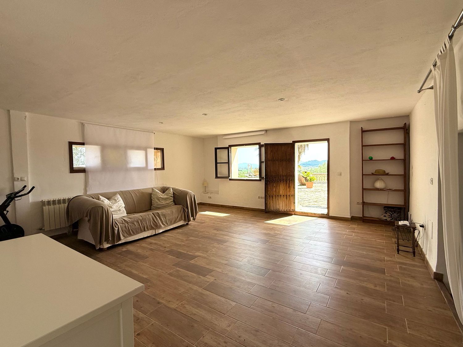Spacious living room with wood flooring, neutral walls, sofa, open shelving, and a door leading to a scenic outdoor view.