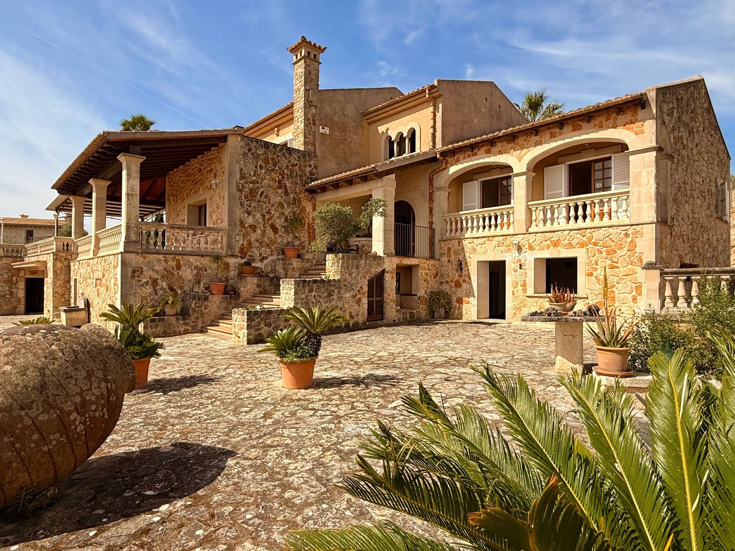 Rustic stone villa with balcony, terracotta pots, and cobblestone courtyard under blue sky.