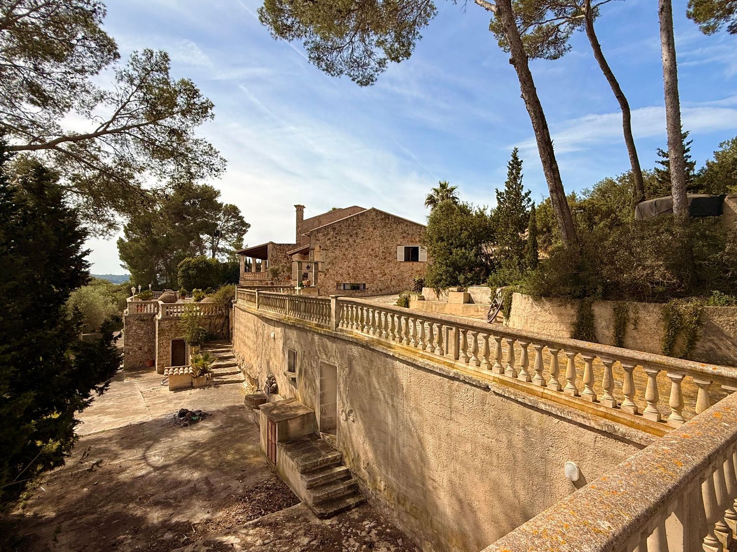 Stone villa with elegant balustrade terrace surrounded by tall pine trees under a blue sky in a Mediterranean setting.
