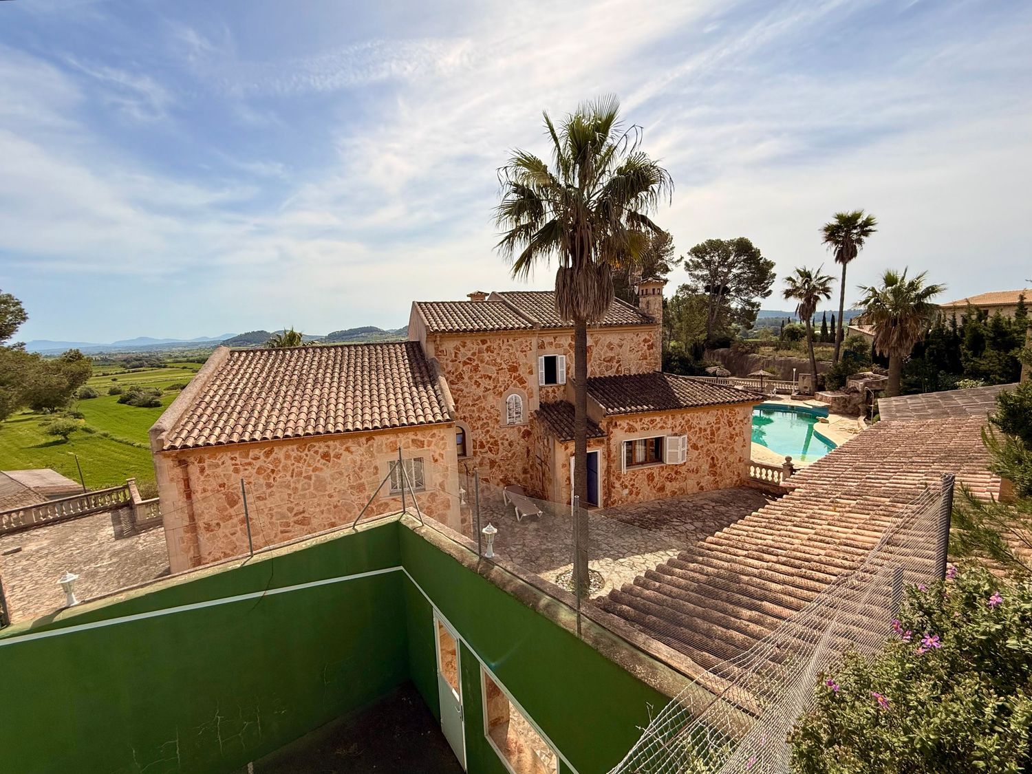 Aerial view of a rustic stone villa with terracotta roof, palm trees, and a swimming pool in a scenic countryside setting.