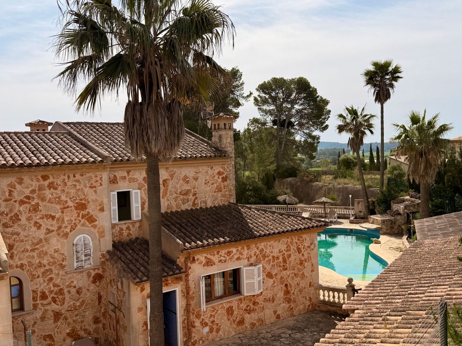 Stone villa with terracotta roof tiles, palm trees, and a blue swimming pool overlooking a scenic Mediterranean landscape.