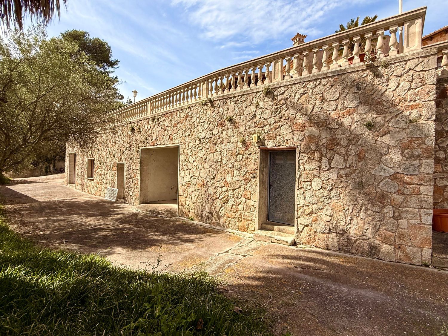 Historic stone building with balustrade terrace surrounded by mature trees on a sunny day.