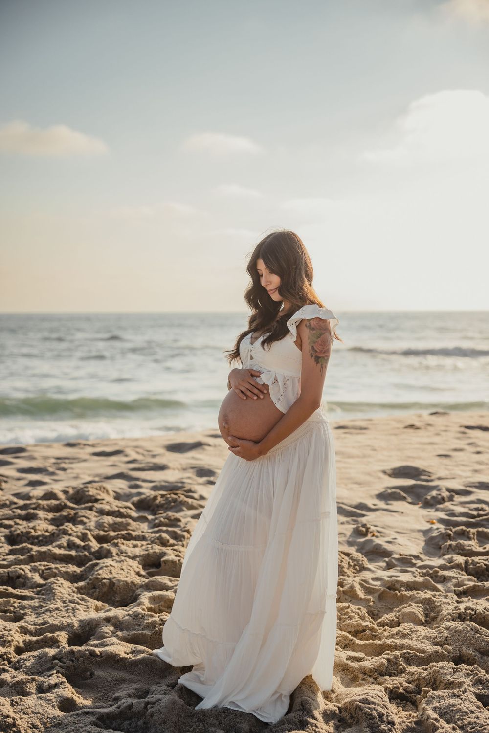 Expectant mother in flowing white dress cradles her baby bump while standing on sandy beach at sunset.