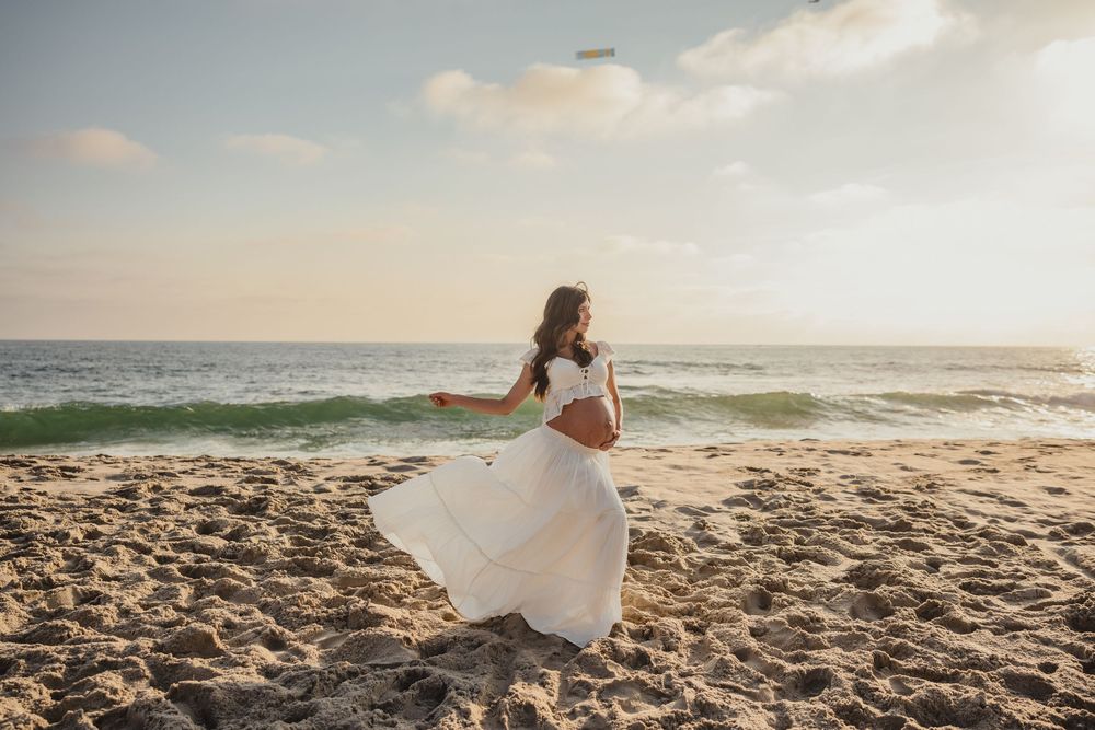 A flowing white dress twirls on a sandy beach at sunset as waves crash along the shoreline.