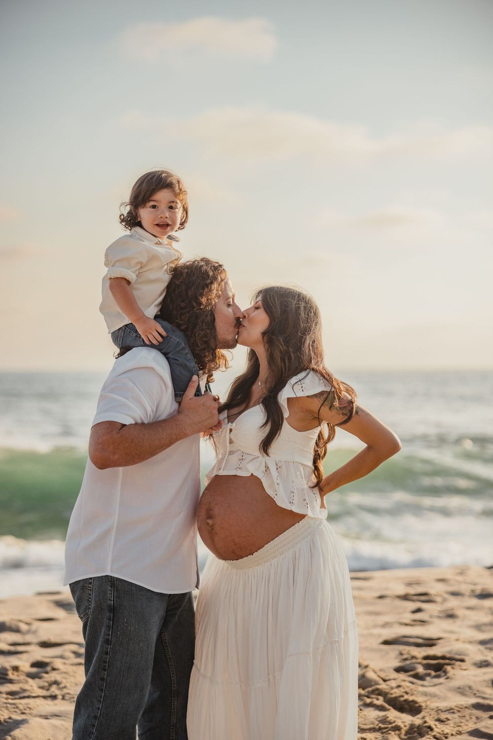 A family shares a tender moment together on the beach at sunset, with waves crashing behind them.