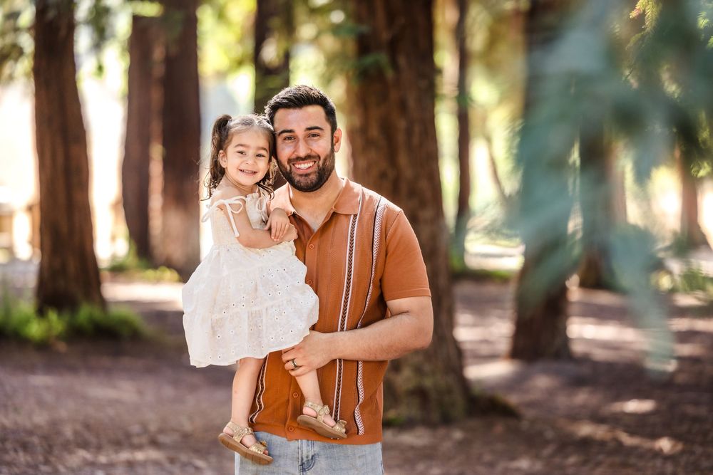 Two people share a joyful moment together while standing in a sunlit forest path with tall trees in the background.