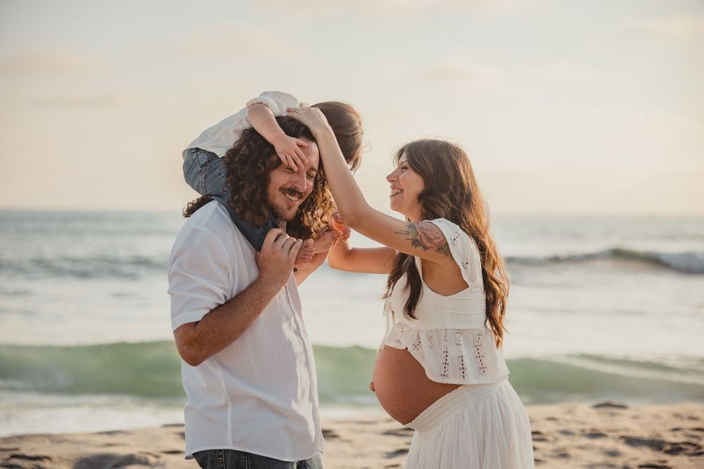 A playful couple laughs together at sunset on a sandy beach with ocean waves in the background.