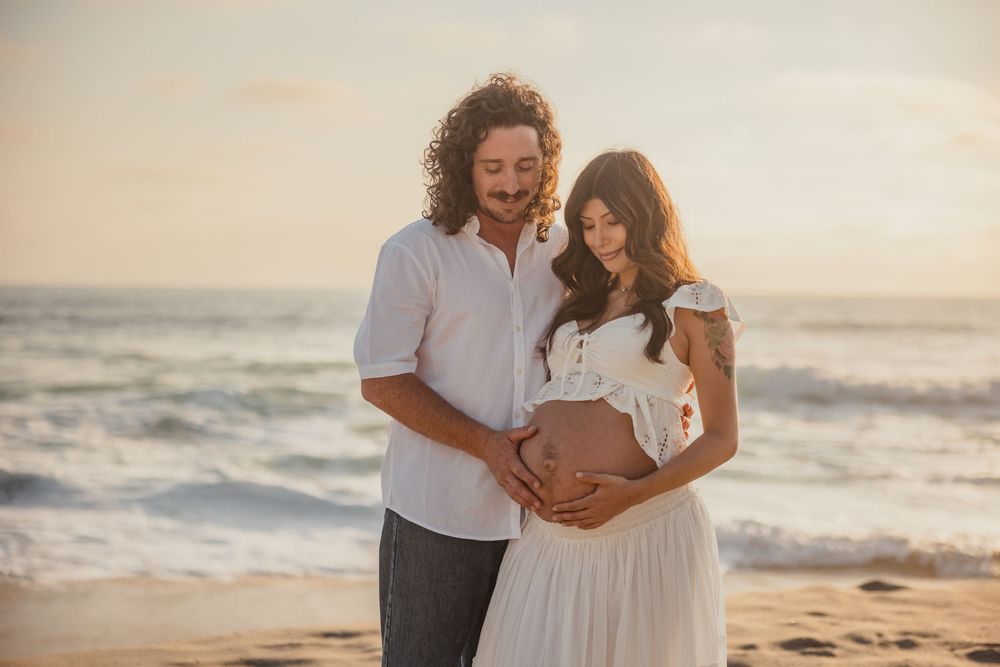 A couple stands on a beach at sunset, with the person in white wearing a flowing dress and touching their belly.