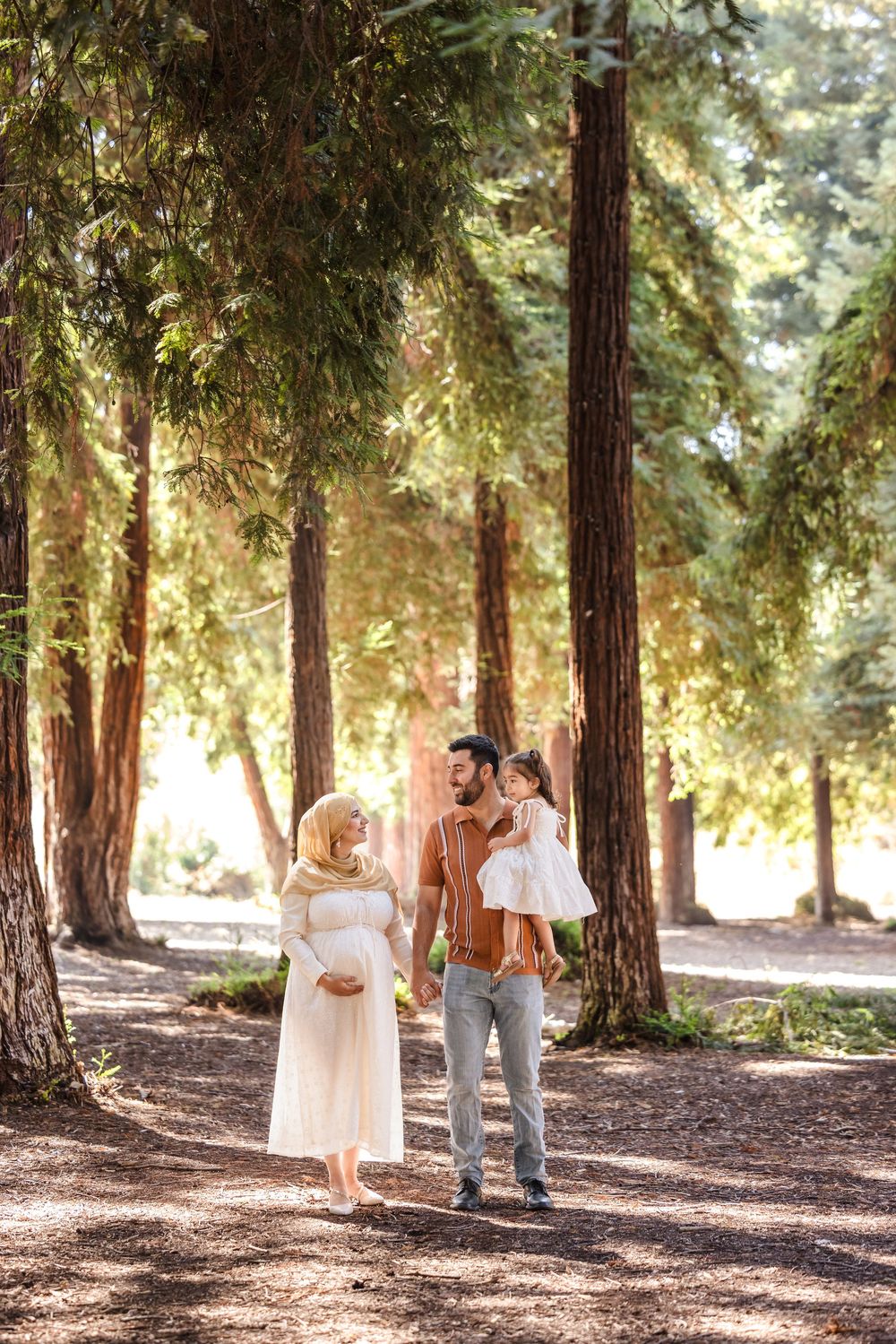 A family walks along a sunlit path through towering redwood trees in a serene forest setting.