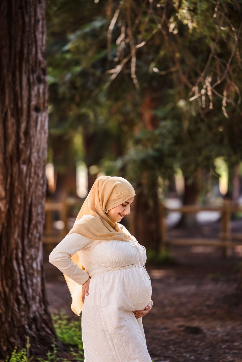 A pregnant woman in a white dress and beige hijab stands in a peaceful woodland setting during golden hour.