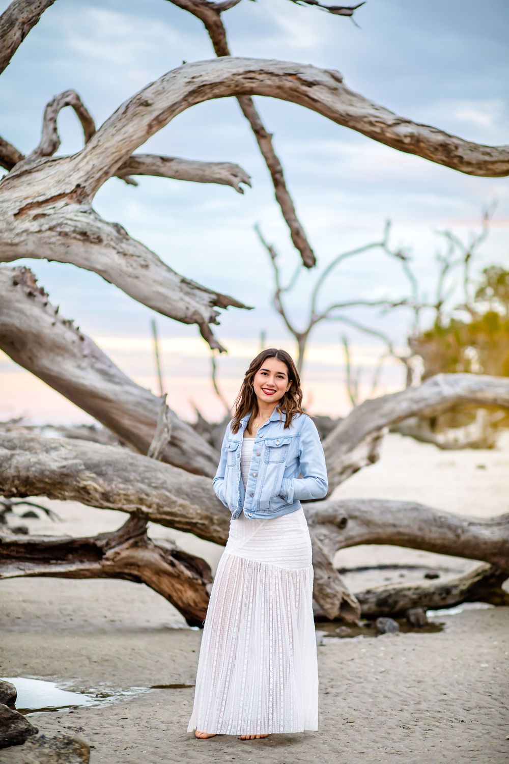 A white pleated maxi skirt and denim jacket outfit photographed against weathered driftwood on a beach at sunset.