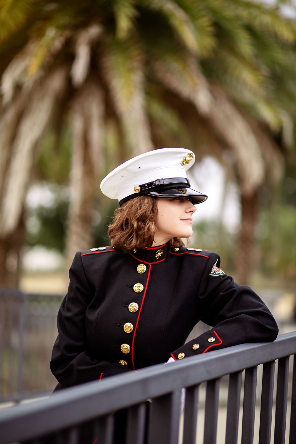 A person in formal Marine dress uniform with white cap and brass buttons leans on a railing with palm trees in background.